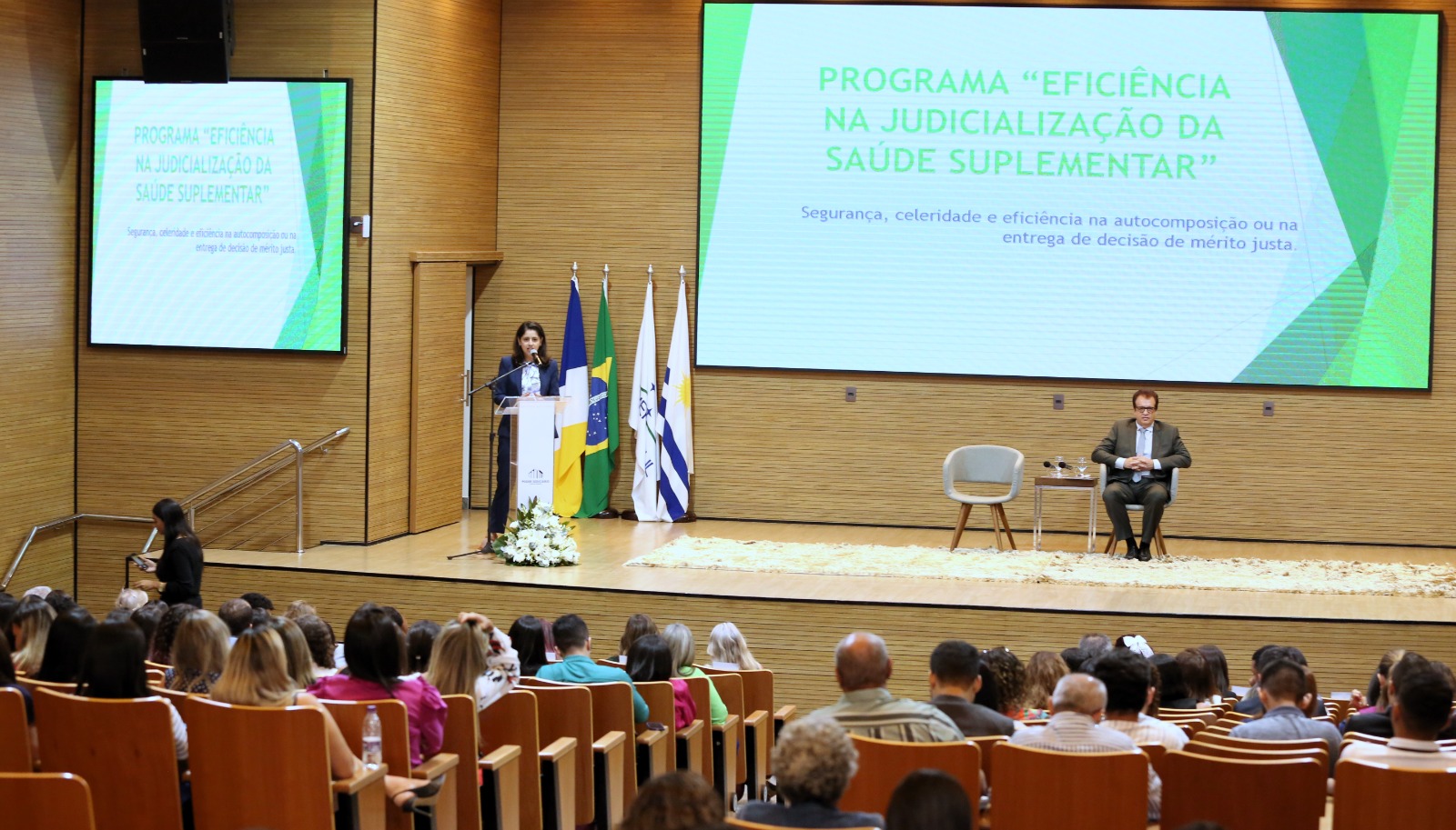 Mulher fala ao microfone em auditório com plateia